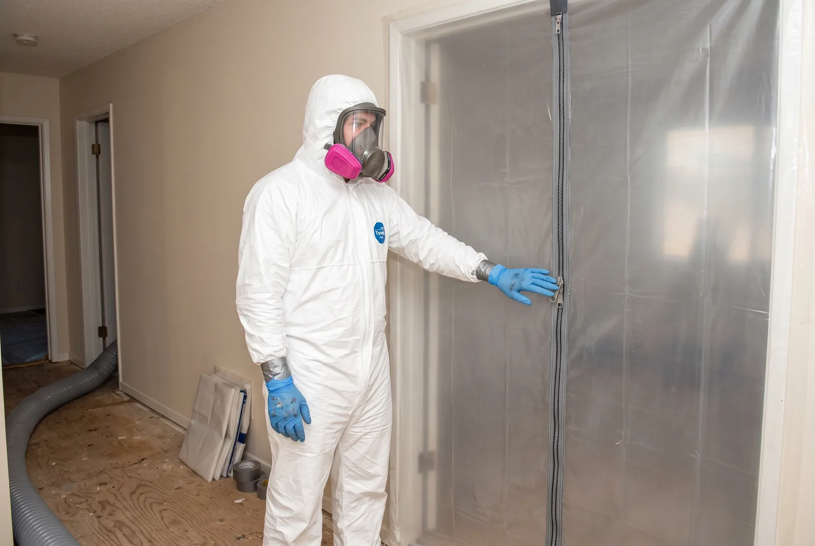 A technician in a sealed white protective coverall and full-face respirator at a zippered plastic containment barrier in a residential hallway, with carpet lifted to bare subfloor and ducting running along the wall.