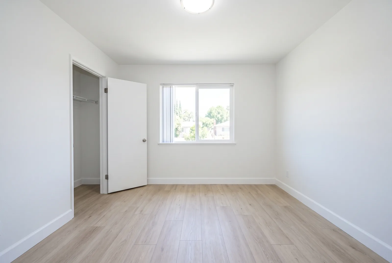 The same bedroom fully restored: fresh white walls, new baseboard, new pale wood flooring, daylight through the window, the room completely empty and clean.
